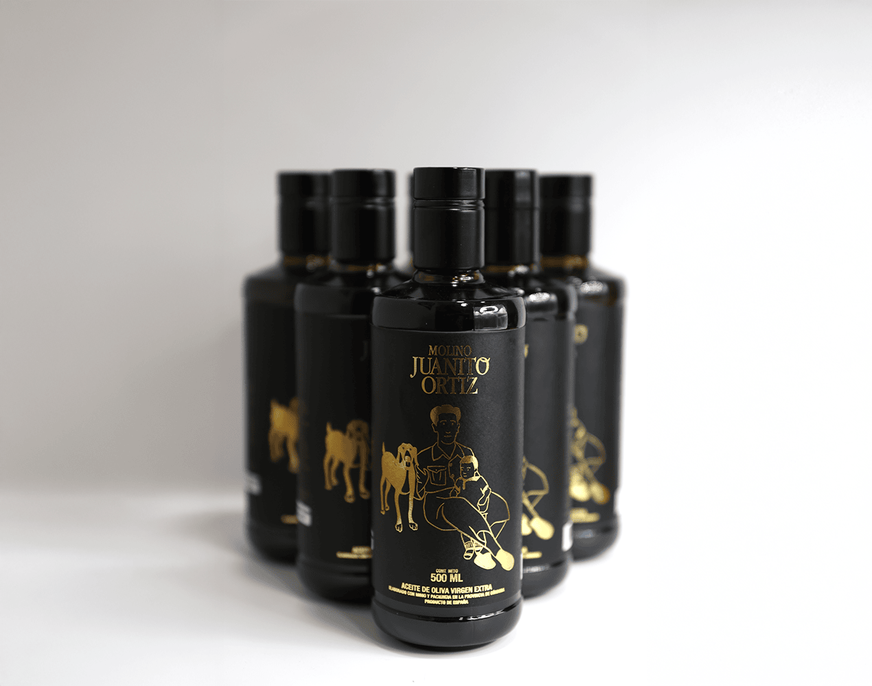 Un paquete de seis botellas de vidrio oscuro de 500 ml de aceite de oliva, de la marca Molino Juanito Ortiz. Cada botella tiene un diseño minimalista en dorado que muestra a un hombre sentado con un perro y un niño, y el logotipo de la marca. El fondo es liso y de color claro.