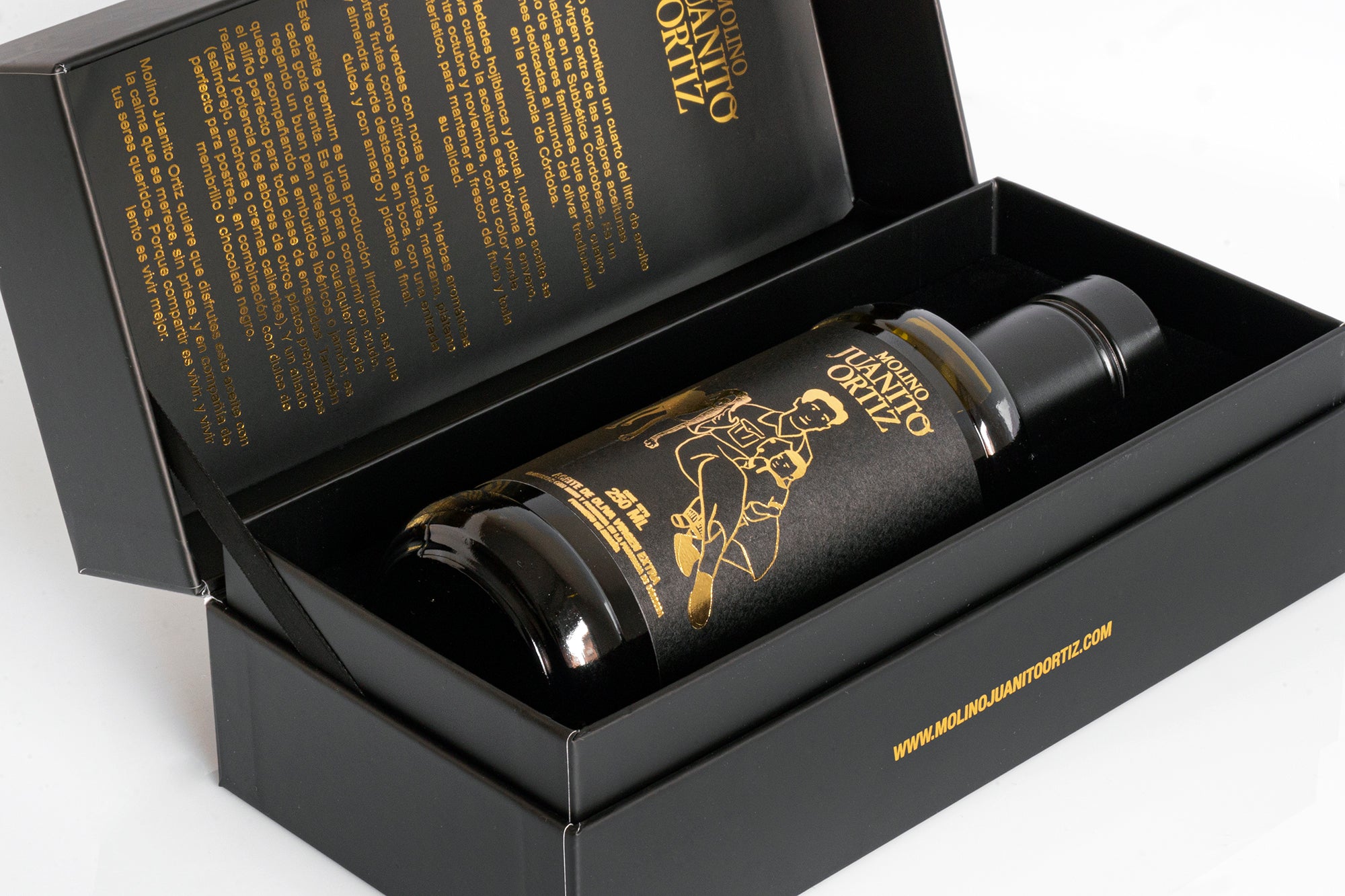 Una botella de aceite de oliva "Molino Juanito Ortiz" con su diseño dorado, colocada dentro de una caja de regalo negra abierta. El interior de la tapa muestra una descripción del producto en texto dorado.