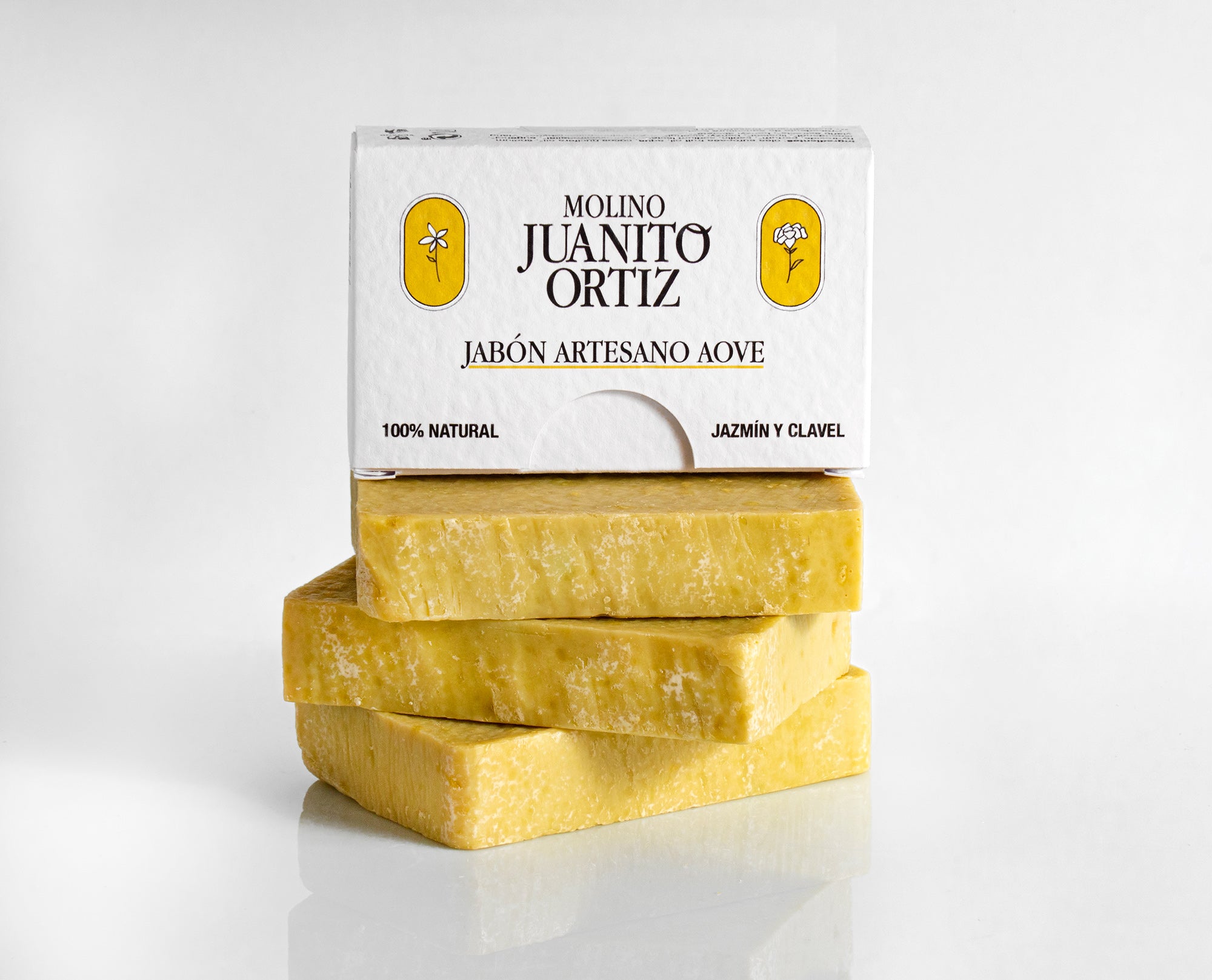 Tres barras de jabón artesanal de color amarillo claro apiladas verticalmente, con una caja de cartón blanca con el texto "MOLINO JUANITO ORTIZ JABÓN ARTESANO AOVE" y las flores de jazmín y clavel encima.