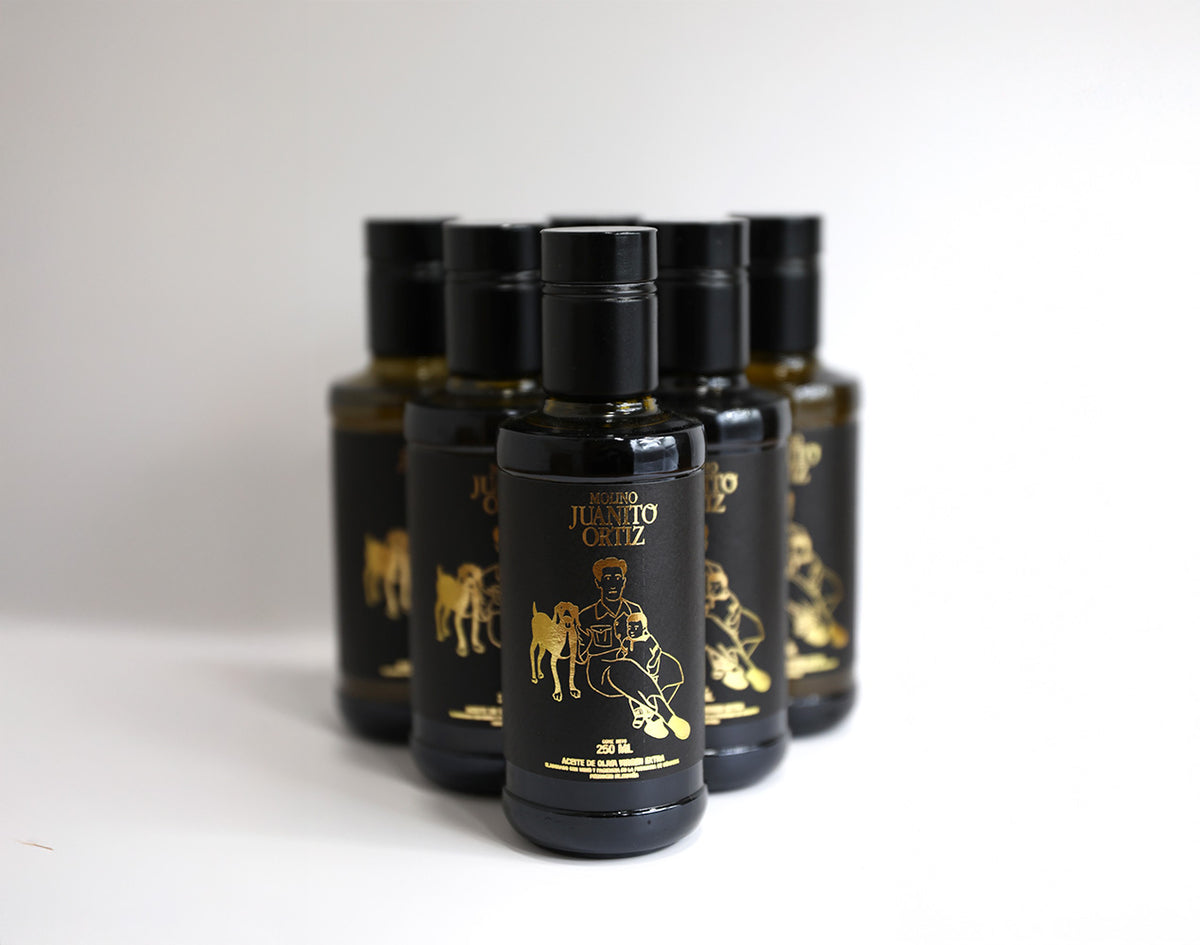 "Un paquete de seis botellas de vidrio oscuro de 250 ml de aceite de oliva, de la marca Molino Juanito Ortiz. Cada botella tiene un diseño minimalista en dorado que muestra a un hombre sentado con un perro y un niño, y el logotipo de la marca. El fondo es liso y de color claro.