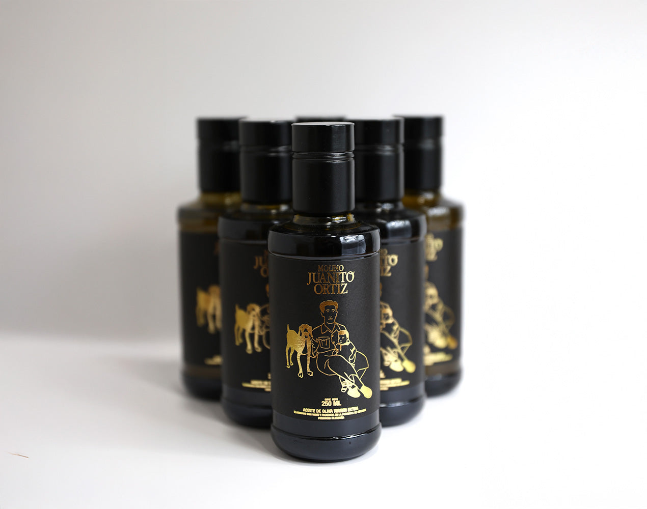 "Un paquete de seis botellas de vidrio oscuro de 250 ml de aceite de oliva, de la marca Molino Juanito Ortiz. Cada botella tiene un diseño minimalista en dorado que muestra a un hombre sentado con un perro y un niño, y el logotipo de la marca. El fondo es liso y de color claro.