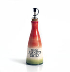 Una aceitera de cerámica, alargada y de cuello estrecho, con un tapón de corcho y un dosificador metálico. La aceitera tiene un diseño pintado con franjas degradadas en tonos rojos, rosados y verdes, con el logotipo "Molino Juanito Ortiz" en negro. El fondo es liso y de color claro.