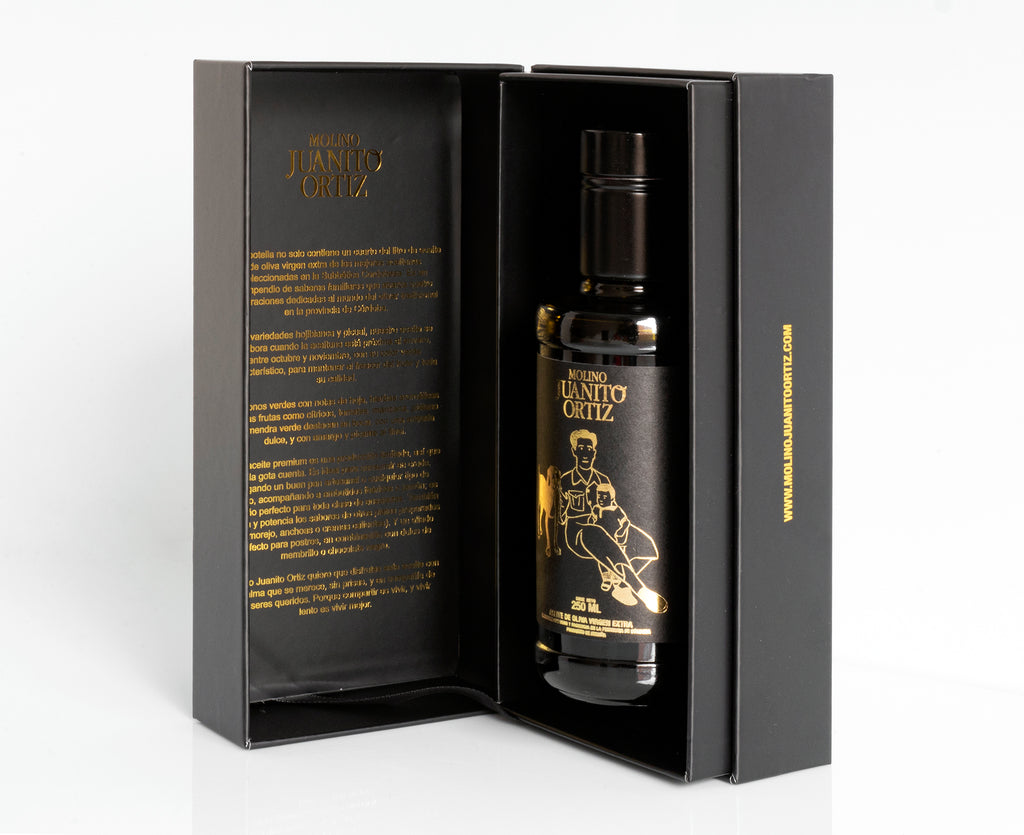  Una vista de la botella de aceite de oliva "Molino Juanito Ortiz" dentro de su caja de regalo negra abierta, mostrando el diseño dorado de la botella y la información del producto impresa en el interior de la tapa.