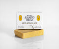 Una barra de jabón artesanal de color amarillo claro colocada horizontalmente sobre una superficie reflectante, con una caja de cartón blanca con el texto "MOLINO JUANITO ORTIZ JABÓN ARTESANO AOVE" y las flores de jazmín y clavel encima.