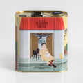 Una lata cuadrada de aceite de oliva "Molino Juanito Ortiz" con la ilustración delantera que muestra a un hombre sentado con un bebé y un perro frente a una puerta.